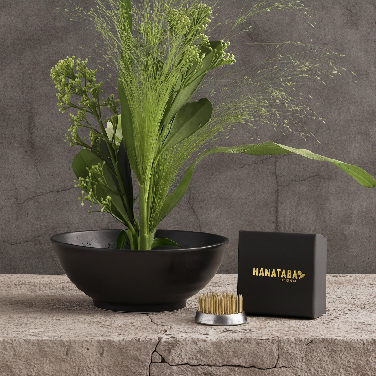 A black ceramic bowl filled with green reeds and foliage sits on a stone surface beside a black Hanataba box and a small kenzan 34 mm flower frog flower arrangement tool, against a textured gray wall.