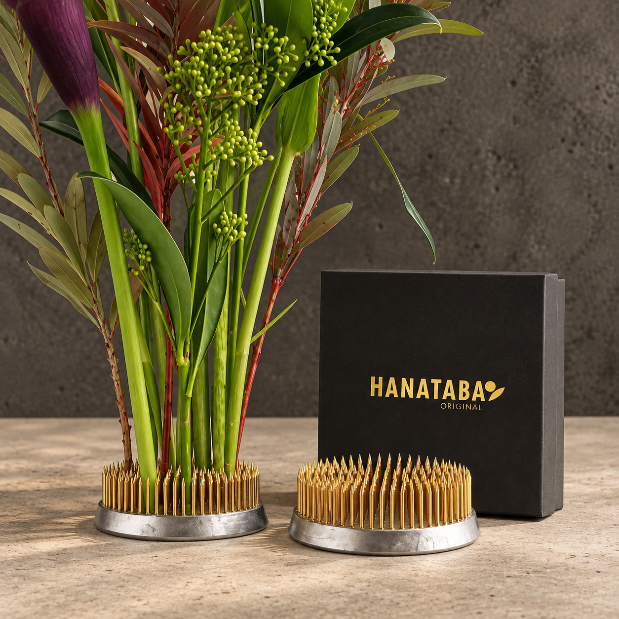 Two Hanataba Original Kenzan 70 mm displayed on a stone surface, with one standing upright in a floral arrangement and the other lying flat. A black Hanataba Original box is positioned behind them against a textured gray wall.