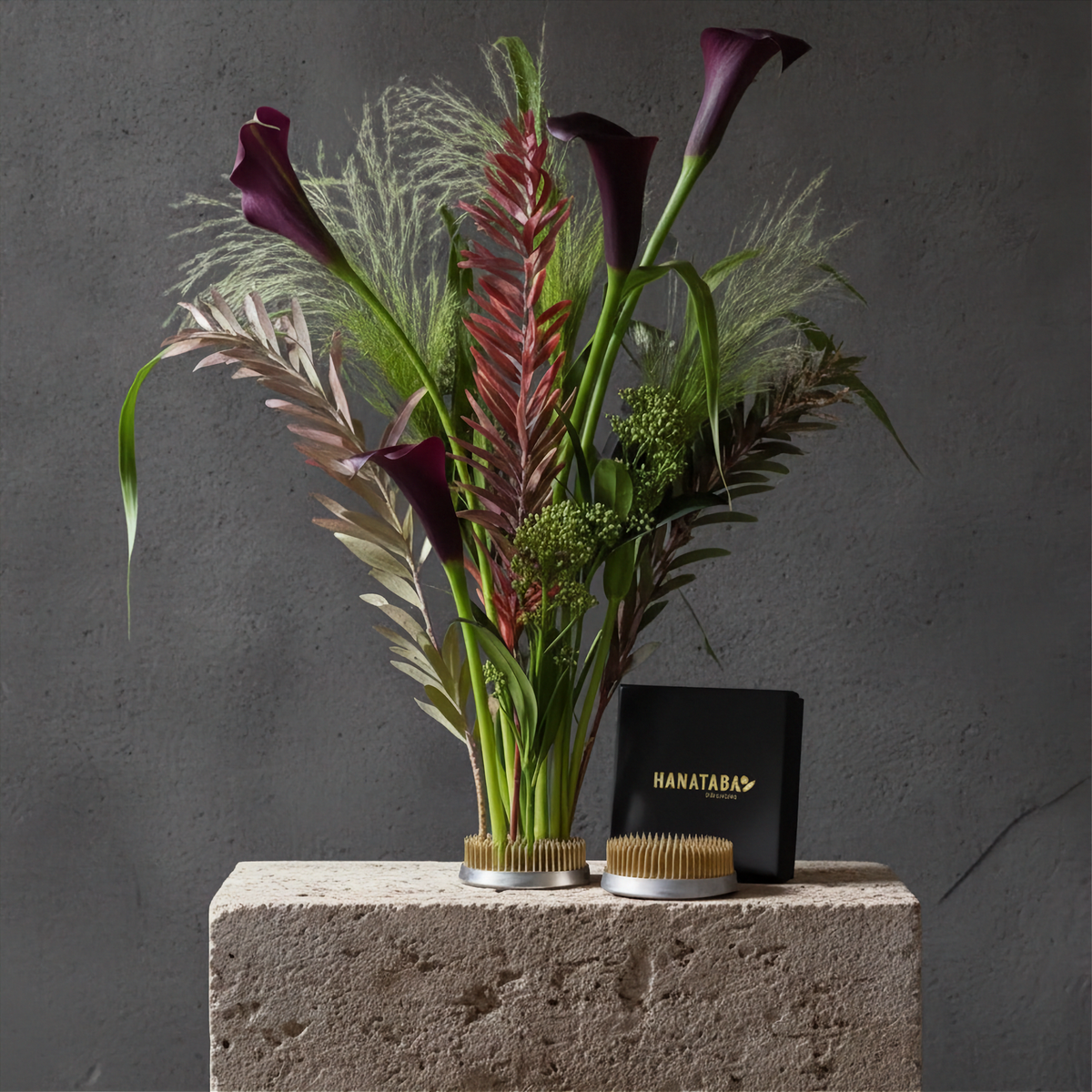 A styled floral arrangement in a Hanataba Original Kenzan 70 mm flower frog pin holder with dark purple calla lilies, green foliage, and feathery filler stems, displayed on a stone block next to a black Hanatara box and two gold-handled brushes against a dark gray wall.