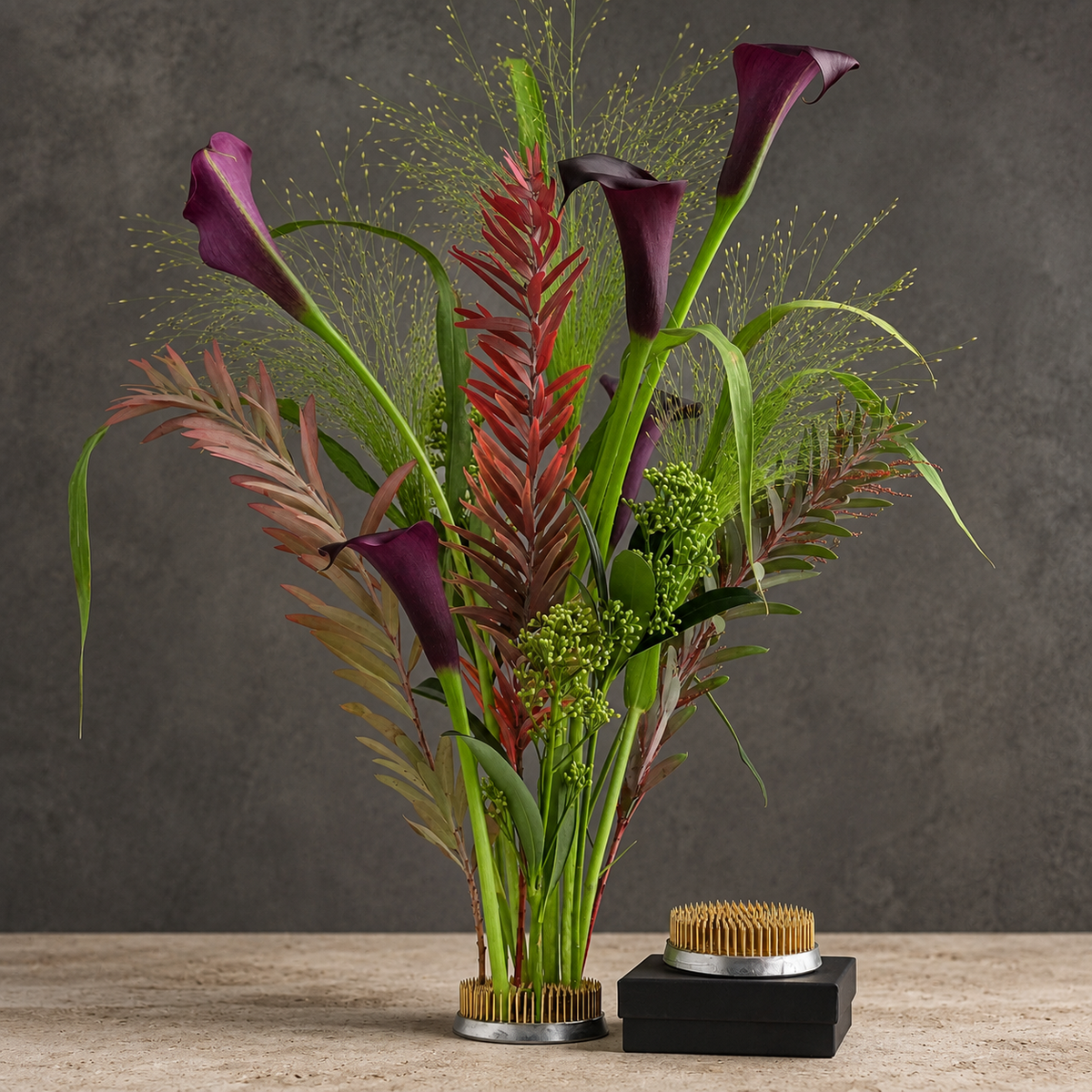 A floral arrangement in a kenzan 70 mm with metal bristle trim, set on a light stone surface against a dark textured wall. The arrangement features dark purple calla lilies, green and reddish-brown foliage, and feathery pale yellow stems.