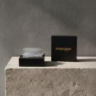 A Hanataba Original Kenzan 70 mm acrylic with a clear ribbed glass bowl on a black base, displayed next to its black branded box on a textured concrete pedestal against a gray wall.
