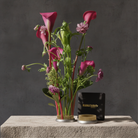A floral arrangement of pink calla lilies and green foliage in a Hanataba Original Kenzan ring 70 mm with gold-toned trim, displayed on a stone pedestal beside a black Hanataba box and a small round gold container against a dark gray textured background.