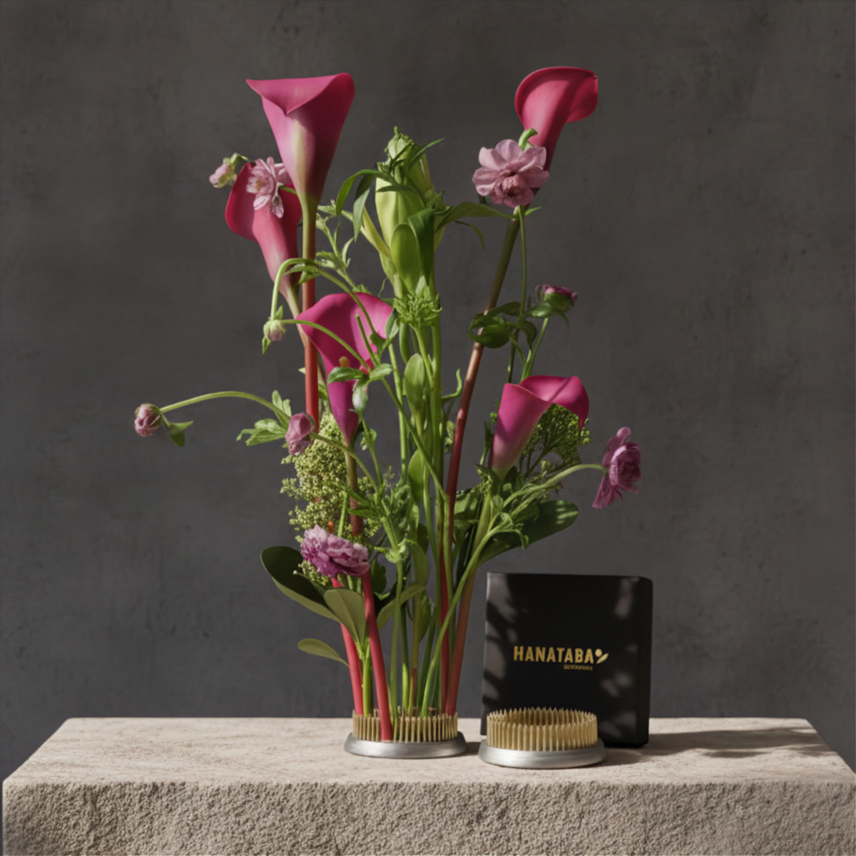 A floral arrangement of pink calla lilies and green foliage in a Hanataba Original Kenzan ring 70 mm with gold-toned trim, displayed on a stone pedestal beside a black Hanataba box and a small round gold container against a dark gray textured background.
