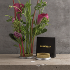A floral arrangement with pink calla lilies and green foliage in a kenzan flower frog pin holder, shown beside a black box labeled HANATABA on a concrete surface against a gray wall.