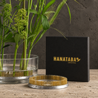 A Hanataba Original Kenzan Ring Flower Frog Ring 120 mm with a black branded box and a glass vase with green floral stems on a stone surface against a dark textured wall.