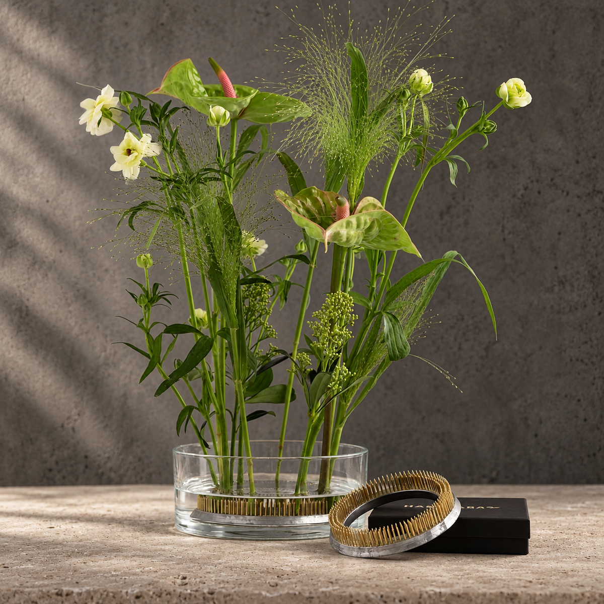 A clear glass vase filled with a mixed floral arrangement of green stems, leaves, and flowers, placed on a kenzan Ring 120 mm Flower Frog. Standing on stone surface against a textured gray wall. A black box and sit beside the vase.
