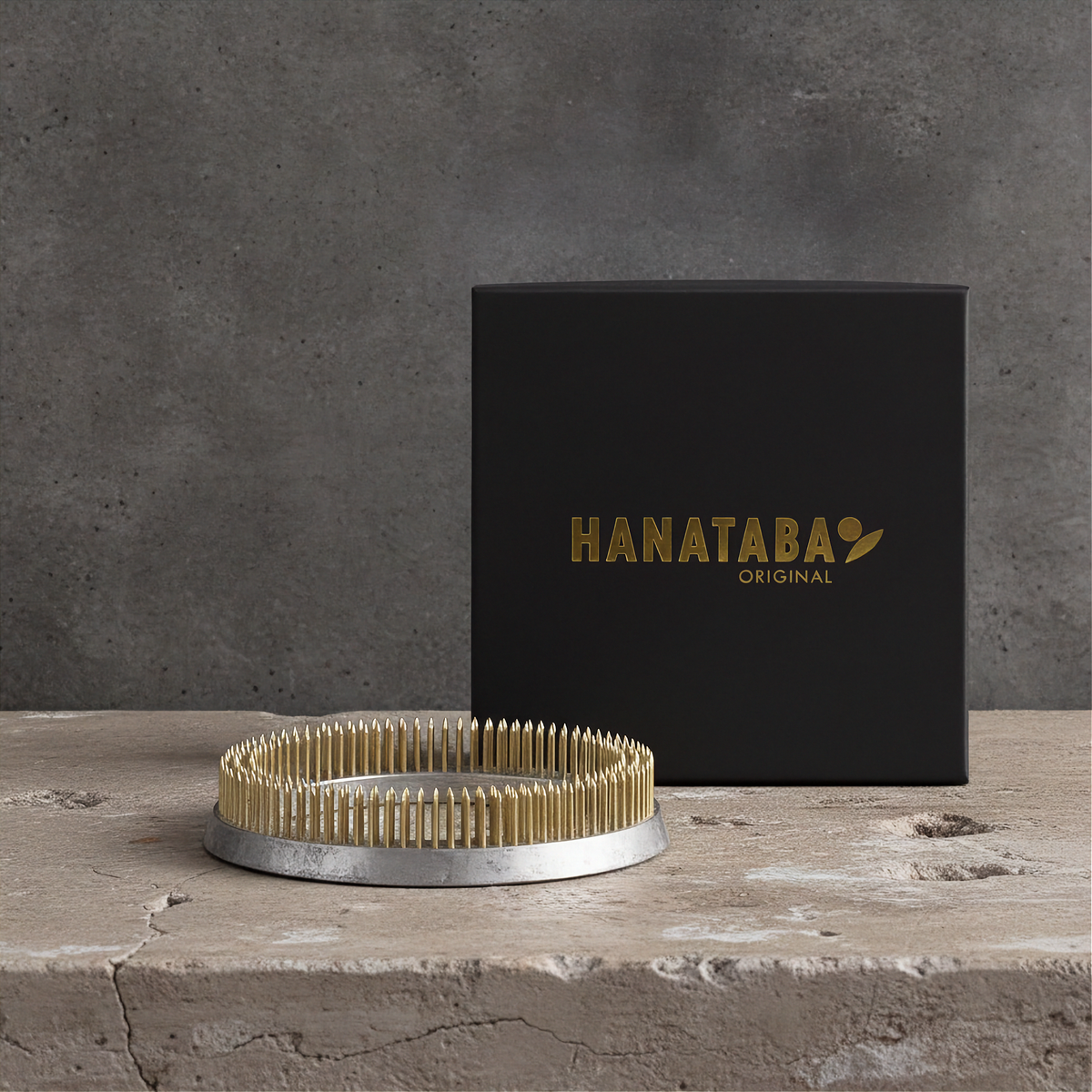 Black box with 'HANATABA' branding and a kenzan ring 120 mm flower frog ring on a concrete block against a gray background. A 120 mm Kenzan flower frog with copper spikes for floral arrangements. The product is shown on top of its black packaging from Hanataba.