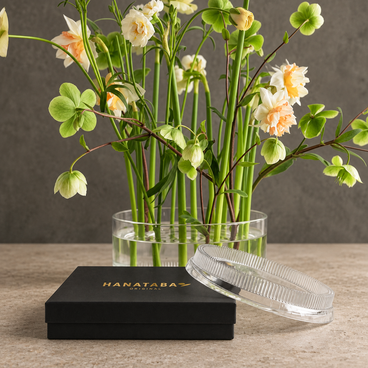 A black Hanataba gift box with gold lettering sits on a stone surface in front of a clear acrylic flower frog kenzan ring 150 mm, filled with green stems and pale flowers. A clear kenzan ring 150 mm acrylic rests beside the box against a neutral gray background.