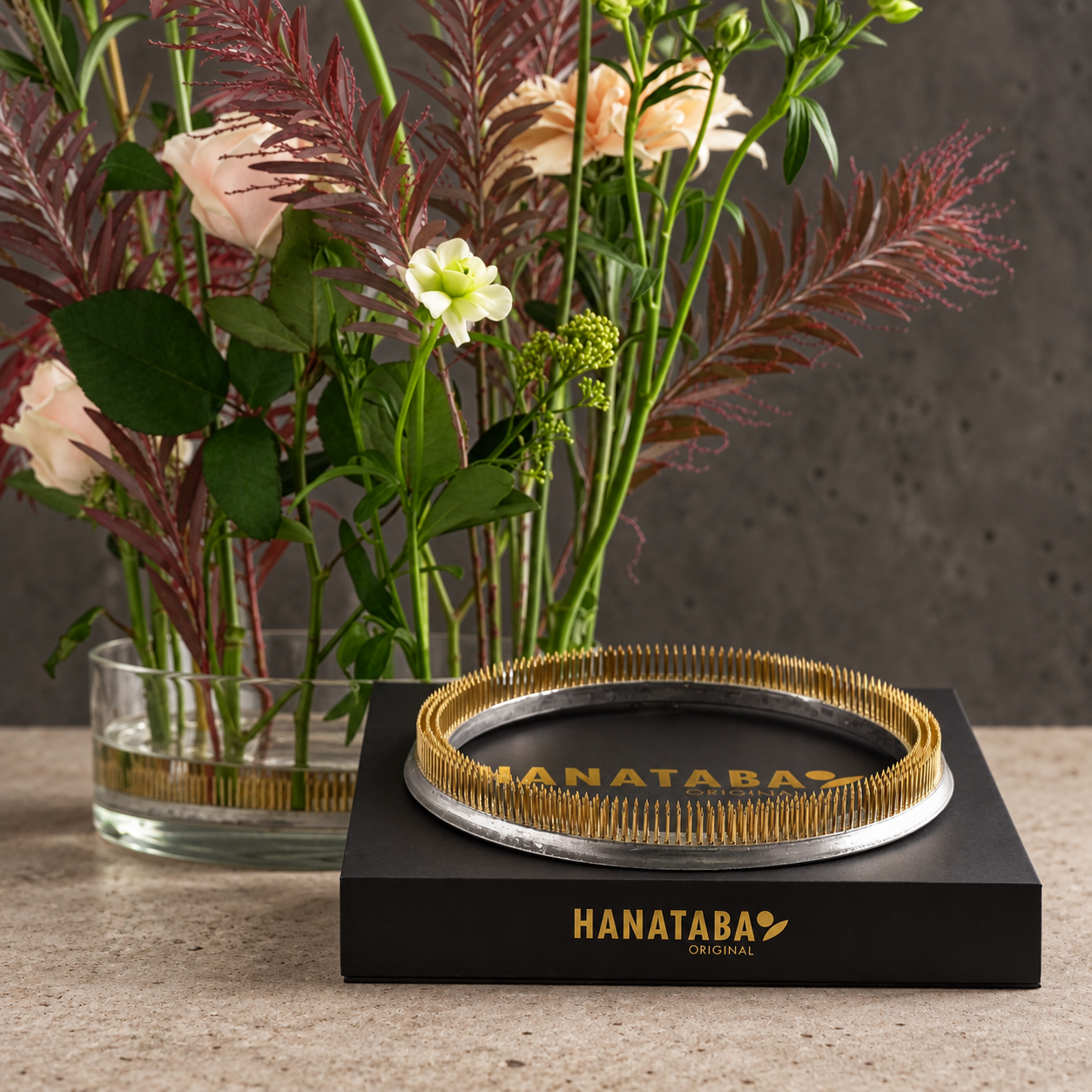 A Hanataba Original flower arrangement in a clear glass vase, styled on a stone surface with a black presentation box in front that reads 'HANATABA ORIGINAL' and a gold Hanataba ring on top.