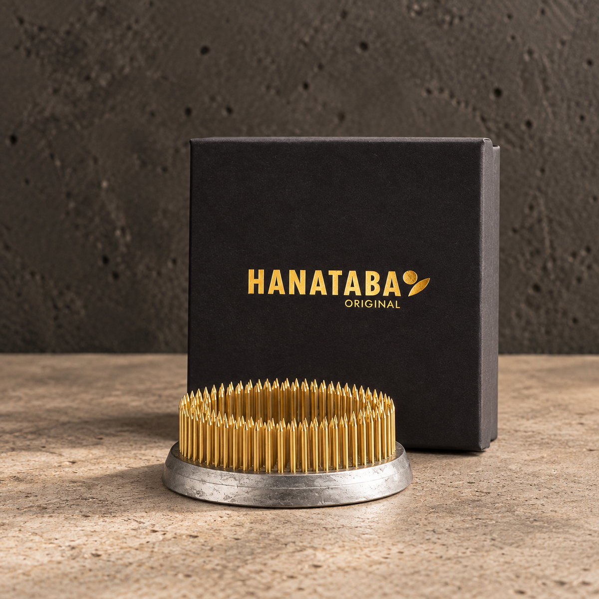 A gold-colored metal Hanataba Original kenzan ring 70 mm with a circular base, displayed in front of its black branded packaging box on a textured stone surface.