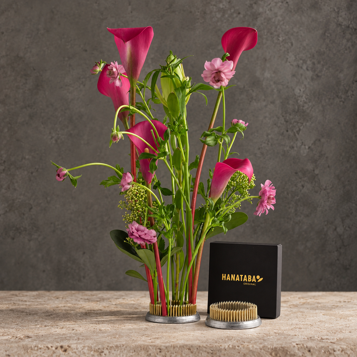 A floral arrangement featuring bright pink calla lilies, pink flowers, and green foliage in HANATABA ORIGINAL kenzan ring 70 mm flower frog pin holder ring with gold-toned bristles at the base, displayed on a stone surface beside a black Hanataba box and a small round container.