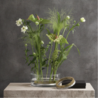 A clear glass vase filled with green and white botanical stems, placed on a light stone pedestal against a textured gray wall. A Hanataba Original Kenzan ring 120 mm and a small black box rest beside the vase.