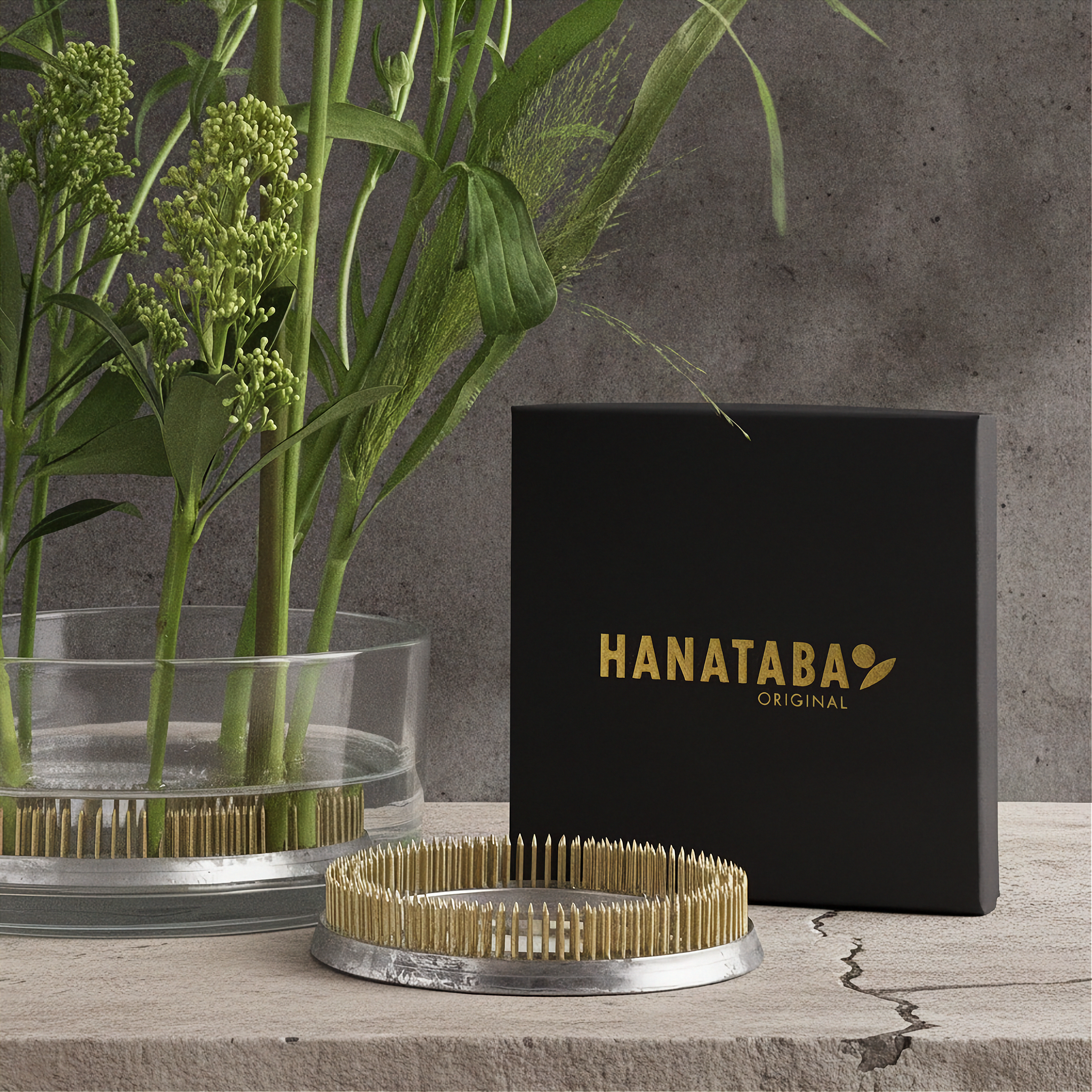 A Hanataba Original product display featuring a black branded box, a round metal tray with light wood bristles, and a glass planter with green stems against a textured gray wall.