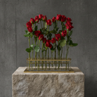 Red roses in a clear vase with gold base on a stone pedestal against a gray concrete wall.