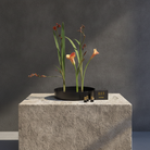 Three small Hanataba Magnetic Mini Kenzan sit on a textured gray stone pedestal next to a black round metal plate containing three orange calla lilies, set against a dark gray textured wall.