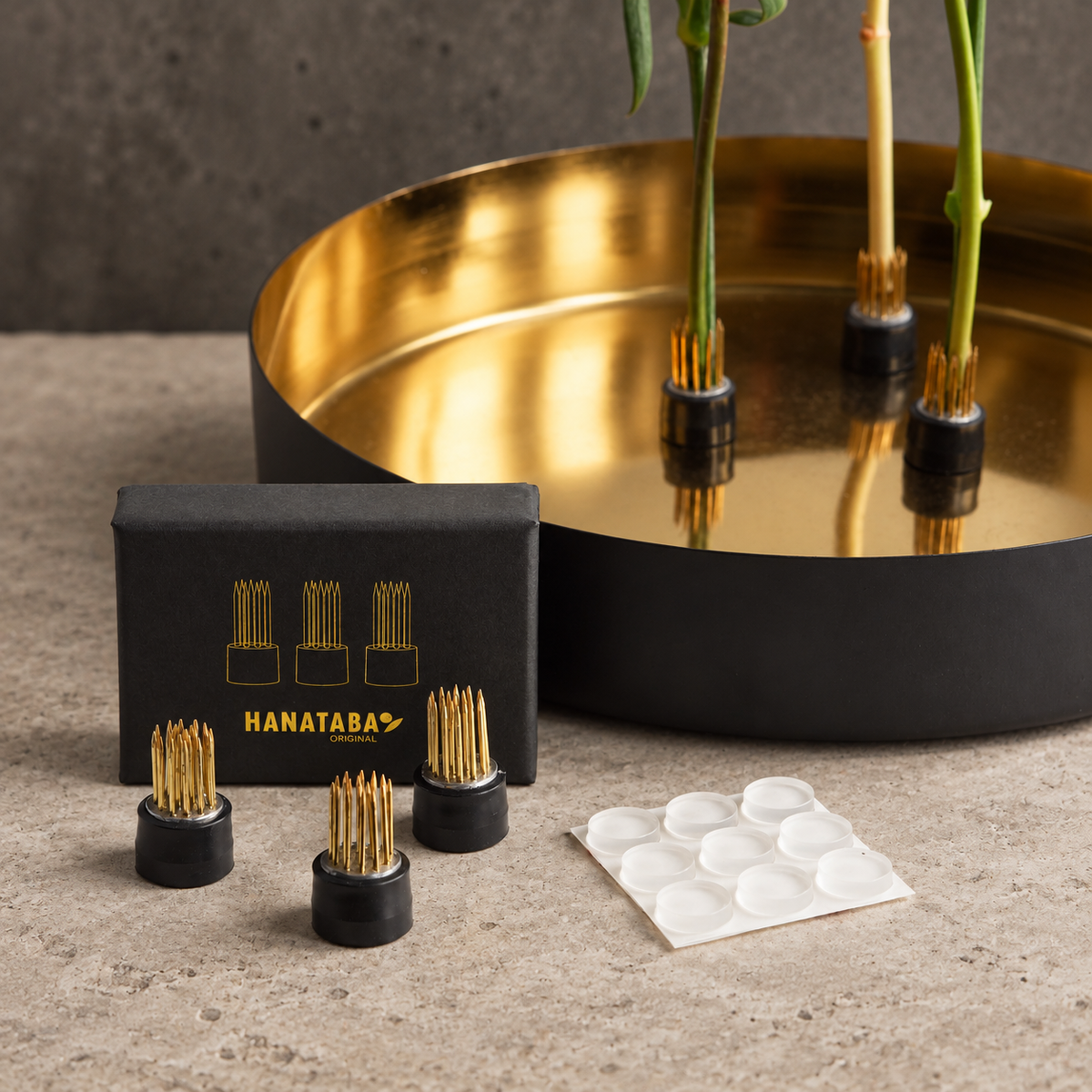 Hanataba Magentic flower frog kenzan with black rubber bases and gold metal needles, shown with a black branded box, a small sheet of white adhesive pads, and a large gold-and-black tray holding several plants.