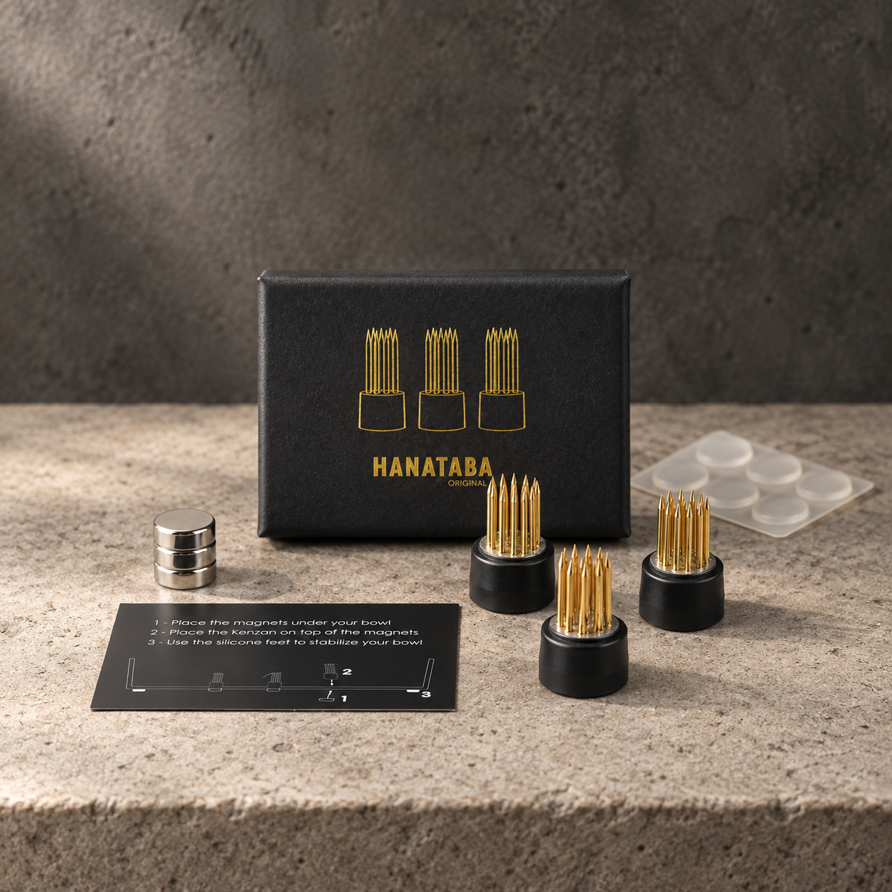 Hanataba Magnetic Mini Kenzan displayed on a stone surface with a dark textured background. The set includes a black presentation box with gold lettering, three black kenzan flower frogs with gold-toned metal pins, three small silver magnets, an instruction card, and a clear plastic tray.