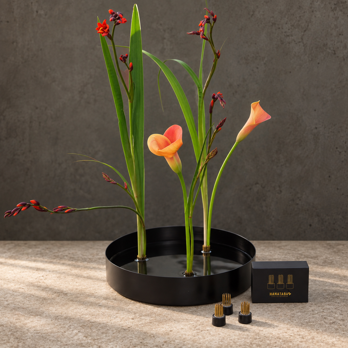 A black round flower arrangement vase in metal. Inside is three magnetic mini kenzan holding three tall green stems with one orange calla lily and several red-orange flower buds, arranged on a stone surface against a textured gray wall. A small black box and three small cylindrical holders are placed in front.