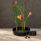 A black round flower arrangement vase in metal. Inside is three magnetic mini kenzan holding three tall green stems with one orange calla lily and several red-orange flower buds, arranged on a stone surface against a textured gray wall. A small black box and three small cylindrical holders are placed in front.