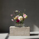 Clear vase with colorful flowers on a concrete block against a dark background