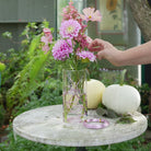 Pink flower arrangement with pink dahlias, arranged in a pink bonsai wire, standing in a transparent glass cylinder vase on a white marble table in an outdoor setting.