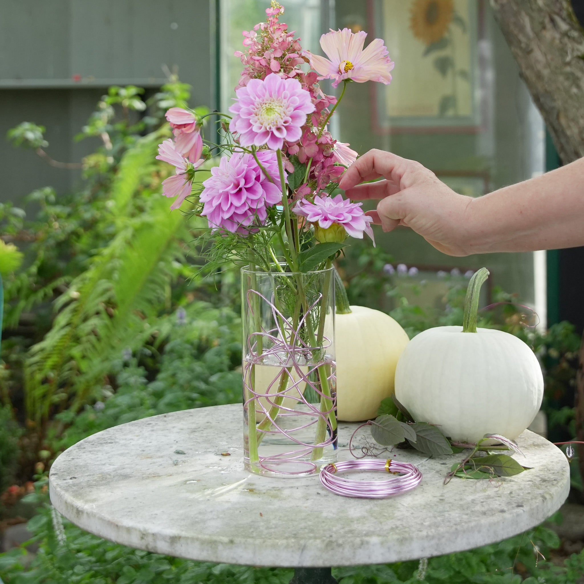 Pink flower arrangement with pink dahlias, arranged in a pink bonsai wire, standing in a transparent glass cylinder vase on a white marble table in an outdoor setting.