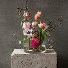 Small glass vase with pink flowers and branches on a concrete block against a gray background