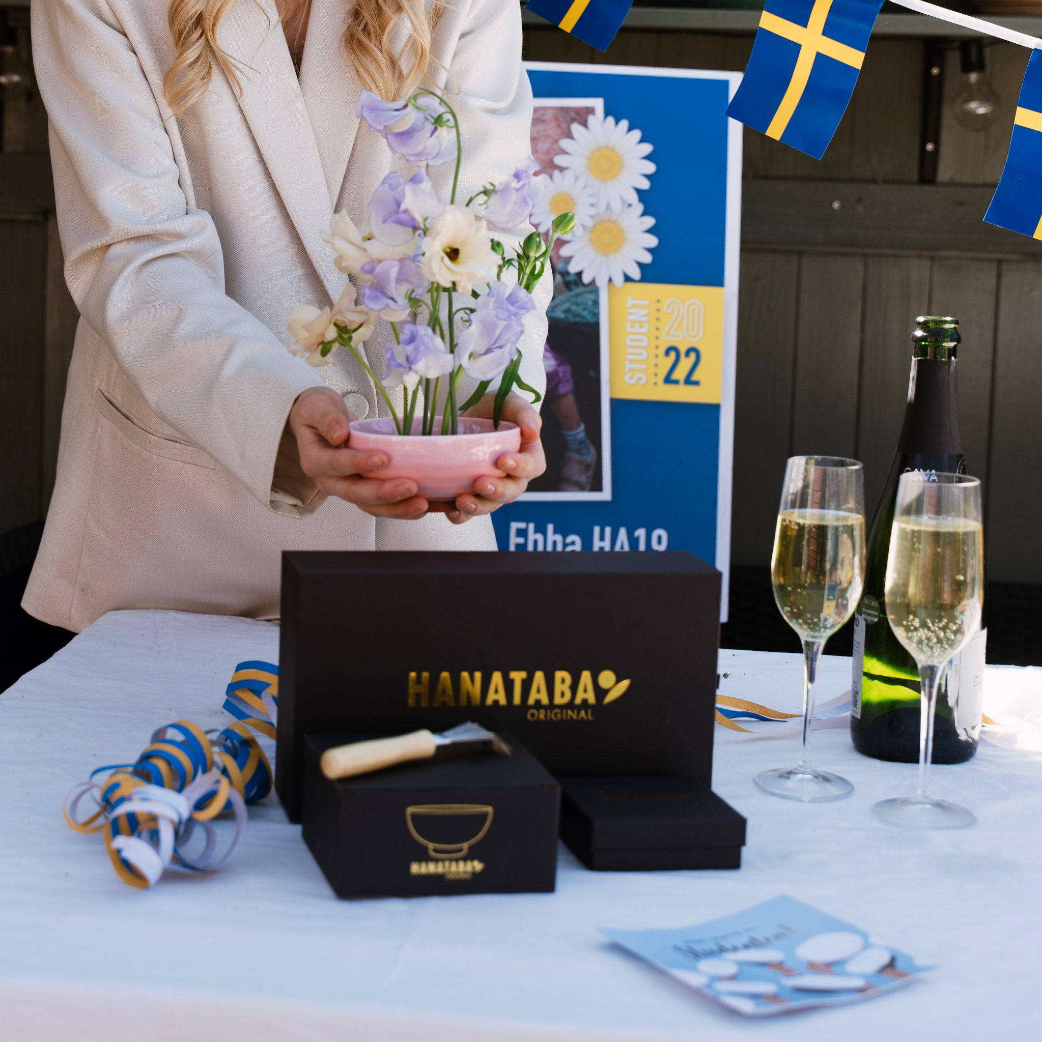 Person holding flowers next to a Hanataba product display with champagne and flags in the background.