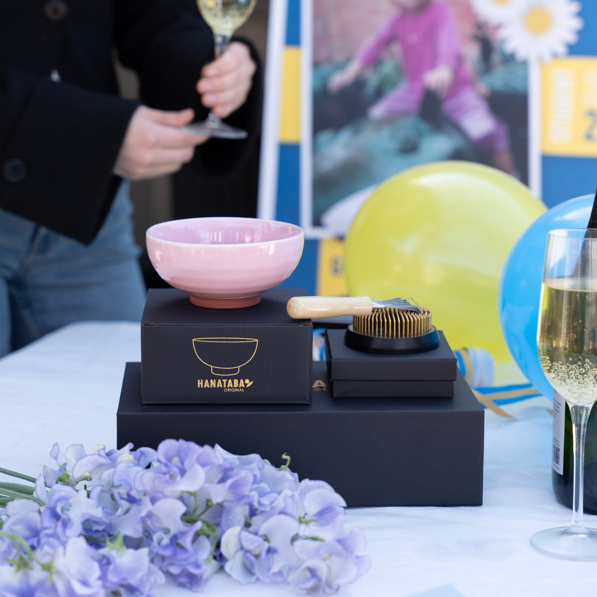 Product display with a pink kenzan 'kobachi' bowl, kenzan brush, kenzan 70 mm and a 'HANATABA' black box on a table with flowers and champagne glasses.
