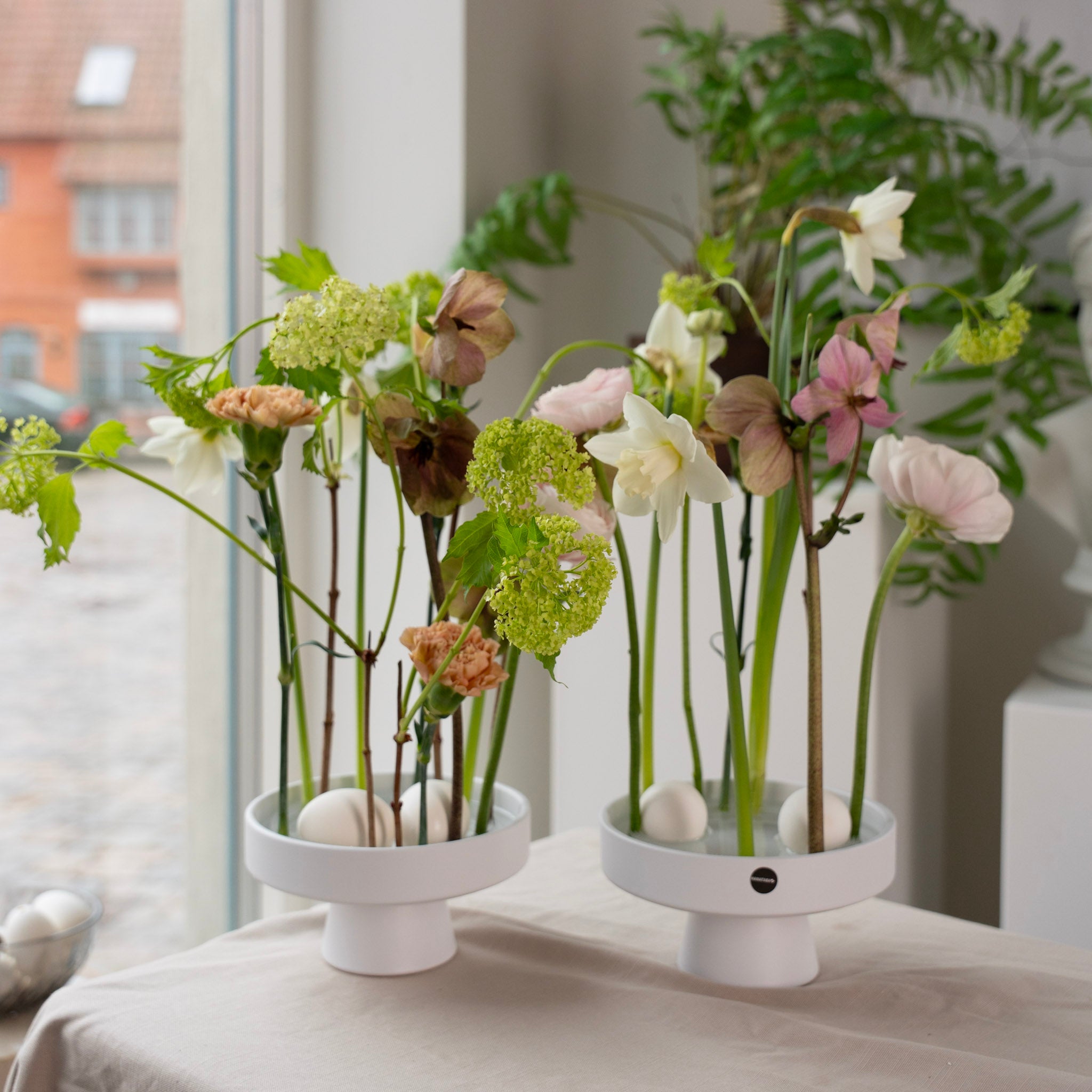 Two white ceramic ikebana vases with floral arrangements used on a hanataba kenzan ring 150mm in transparent acrylic material on a light surface.