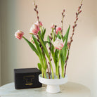 Pink tulips in a white hanataba ceramic ikebana vase on a marble surface with a black box labeled 'Hanataba'.