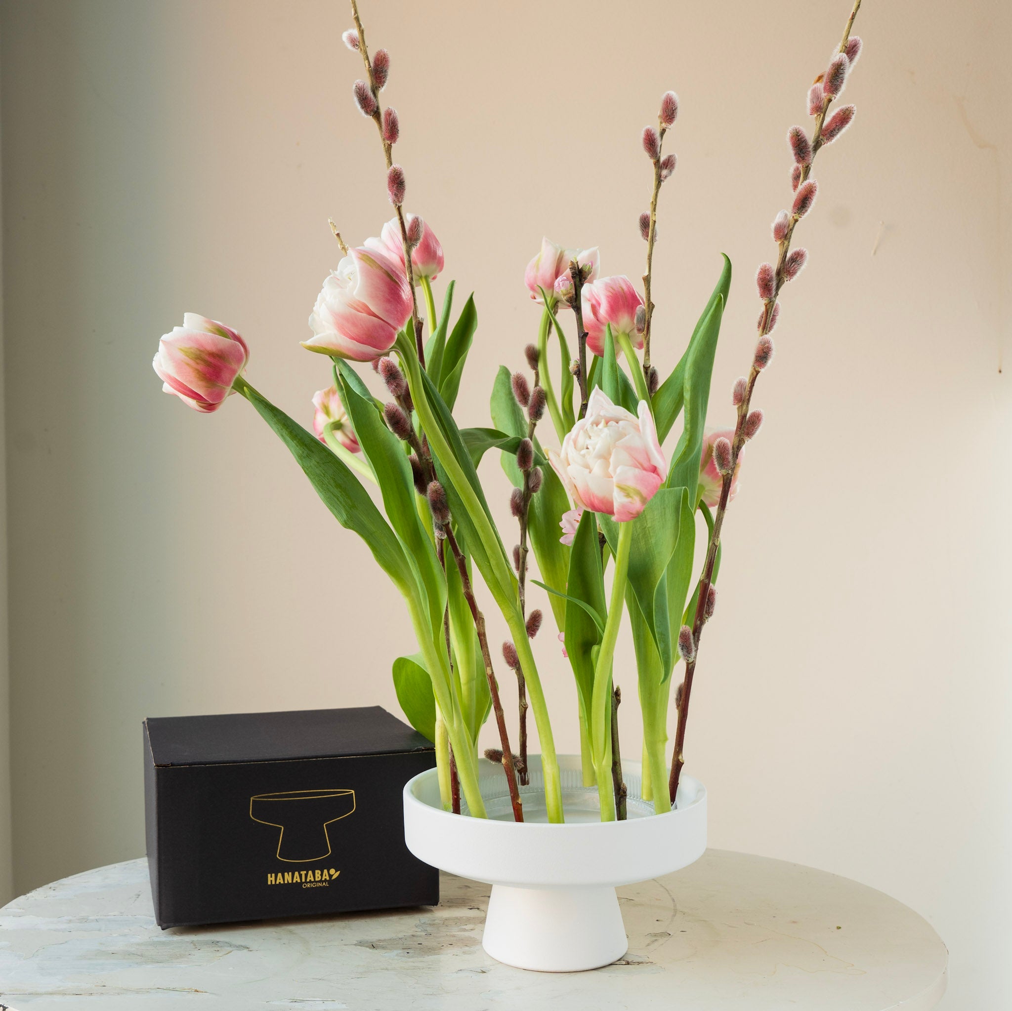 Pink tulips in a white hanataba ceramic ikebana vase on a marble surface with a black box labeled 'Hanataba'.