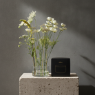 A black square box with gold lettering sits on a textured stone pedestal next to a clear glass vase filled with white and yellow wildflowers, set against a dark gray wall with dramatic shadows.