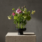 Floral arrangement in a black hanataba ceramic ikebana vase on a concrete block with a dark background
