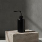 A black plastic bottle with a curved, angled spout, resting on a textured light gray concrete pedestal against a dark gray concrete wall.
