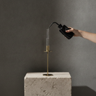 A minimalist brass candle holder with a clear glass vessel, shown on a textured stone pedestal as a hand pours water from a black bottle into the glass.