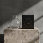 Clear glass vase on a concrete block with a black box featuring a gold logo in the background.