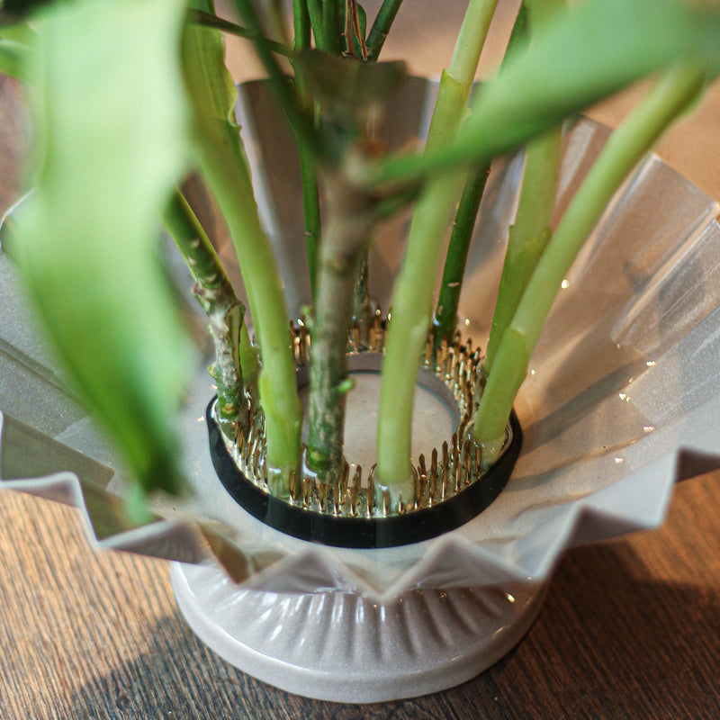 Close-up view of a 70mm kenzan at the base of a flower arrangement, its brass pins securely holding green stems above a rippled glass dish, showcasing the essential tool in ikebana floral art.