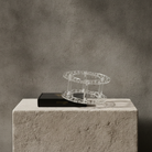 A clear acrylic circular display stand or ring holder placed on top of a black box, set on a textured light stone pedestal against a gray concrete wall.