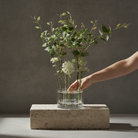 A clear glass vase filled with green eucalyptus stems and small white flowers, placed on a rectangular stone pedestal against a textured gray wall. A person's arm reaches in to adjust the arrangement.