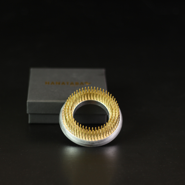 A 70mm kenzan ring with shiny gold-colored needles, perfect for Ikebana floral arrangements, presented beside its dark grey box with 'HANATABA' in gold lettering, on a black surface for contrast