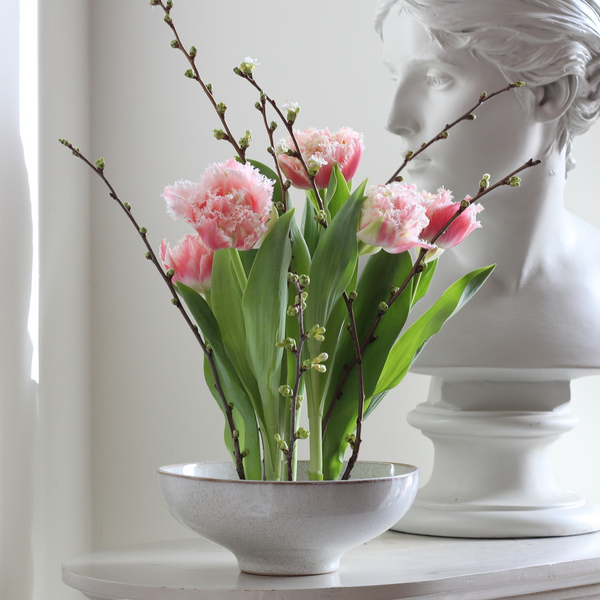 Elegant floral arrangement with pink fringed tulips and delicate budding branches in a speckled bowl, juxtaposed with a classical white bust sculpture.