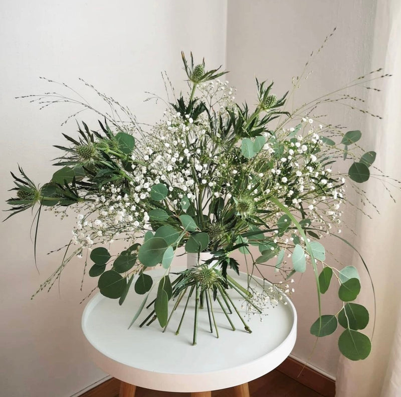 Elegant ikebana arrangement with an assortment of greenery and white blossoms secured by a Hanataba Bouquet Twister in pearly silver on a white table.