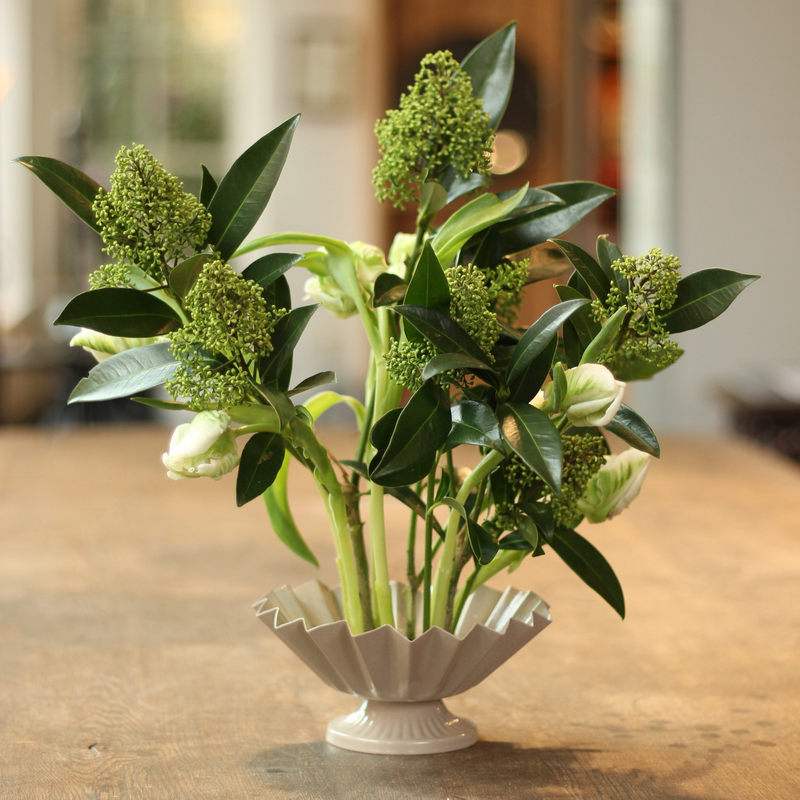 Elegant floral arrangement featuring green budding flowers and lush leaves in a classic white fluted vase anchored on a kenzan ring, presented on a wooden surface with a softly blurred background.
