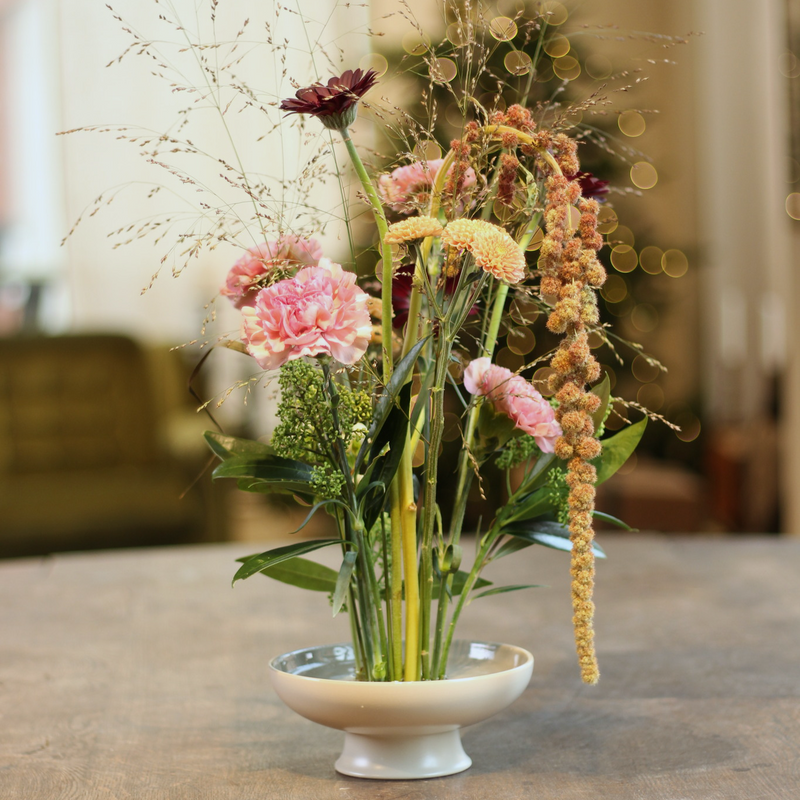 A vibrant arrangement of pink carnations, a deep burgundy dahlia, and delicate hanging amaranths, all anchored on a 70mm kenzan ring, set in a shallow white bowl on a wooden table, invoking a sense of harmony and natural elegance.