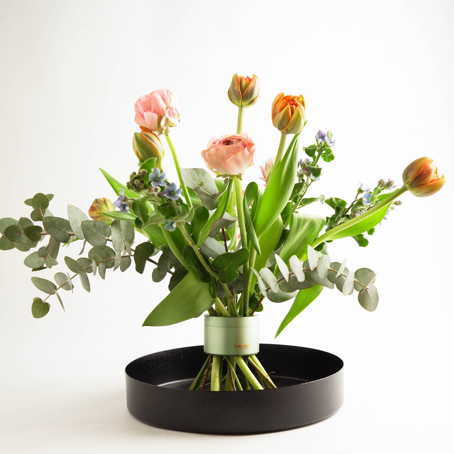 Beautiful bouquet of pink and orange tulips and eucalyptus leaves arranged in a Hanataba Matcha Green Flower Twister that is placed in a black round metal tray vase in a bright white surrounding.