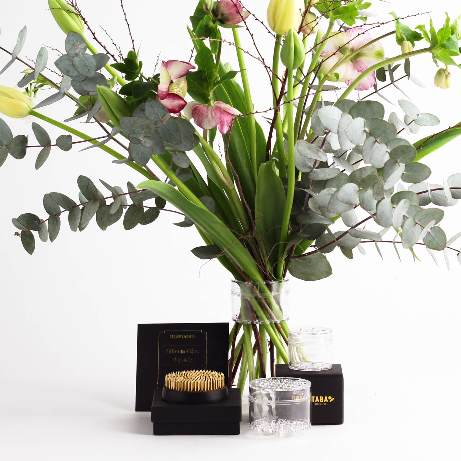 The Hanataba Flower Design Kit displayed with a Crystal Clear Bouquet Twister holding eucalyptus stems, pink-tipped blooms, and green buds, next to a flower pin frog. Elegant black packaging showcases the golden Hanataba logo. All set against a clean white background.