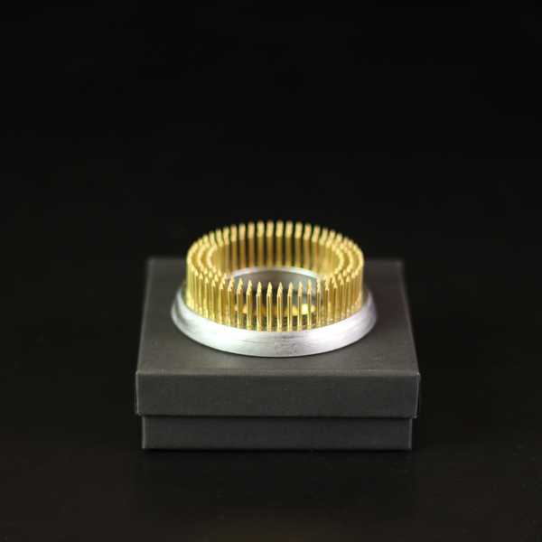 A 70mm kenzan ring with golden pins designed for floral arrangements, displayed on a dark grey box against a black background, highlighting its gleaming metal and precision.
