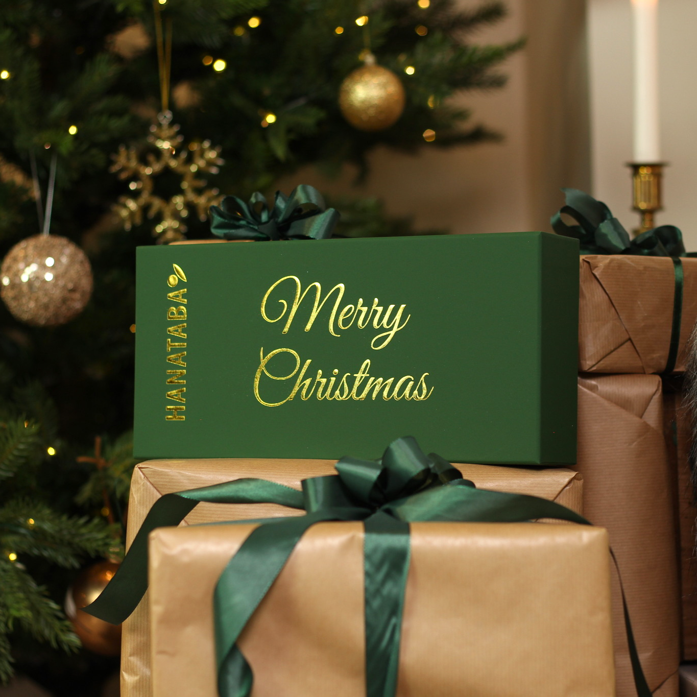 A green Hanataba box with 'Merry Christmas' in gold lettering sits atop a wrapped gift with a green ribbon, against a backdrop of a twinkling Christmas tree.