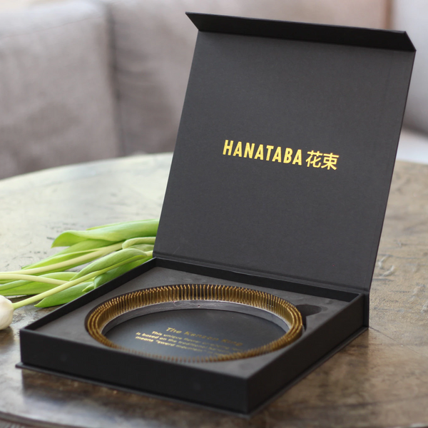 An elegant Hanataba branded black box open to reveal a golden kenzan ring inside, with fresh green tulip stems laid beside it on a textured surface.